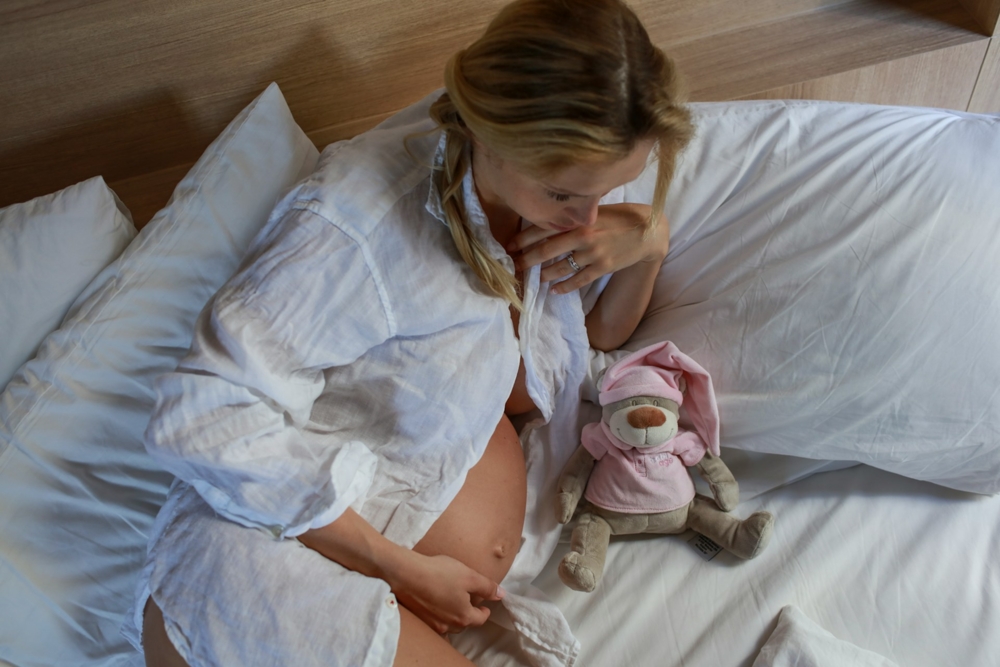 Pregnant woman laying on a bet next to a teddy bear in a pink outfit